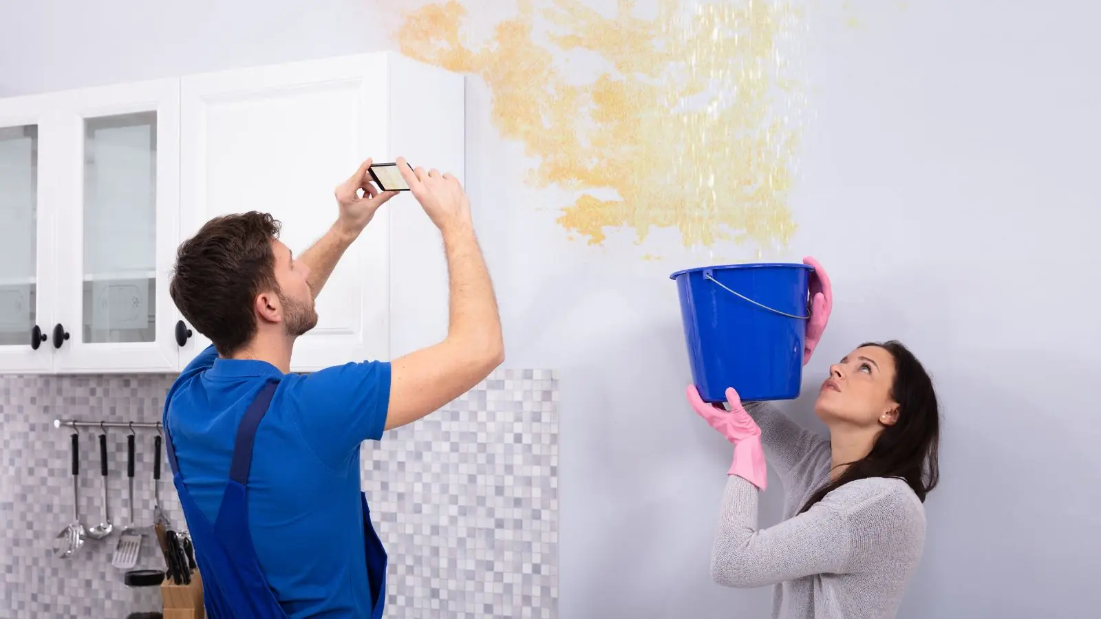Two people photographing a water stain on a wall while one holds a bucket